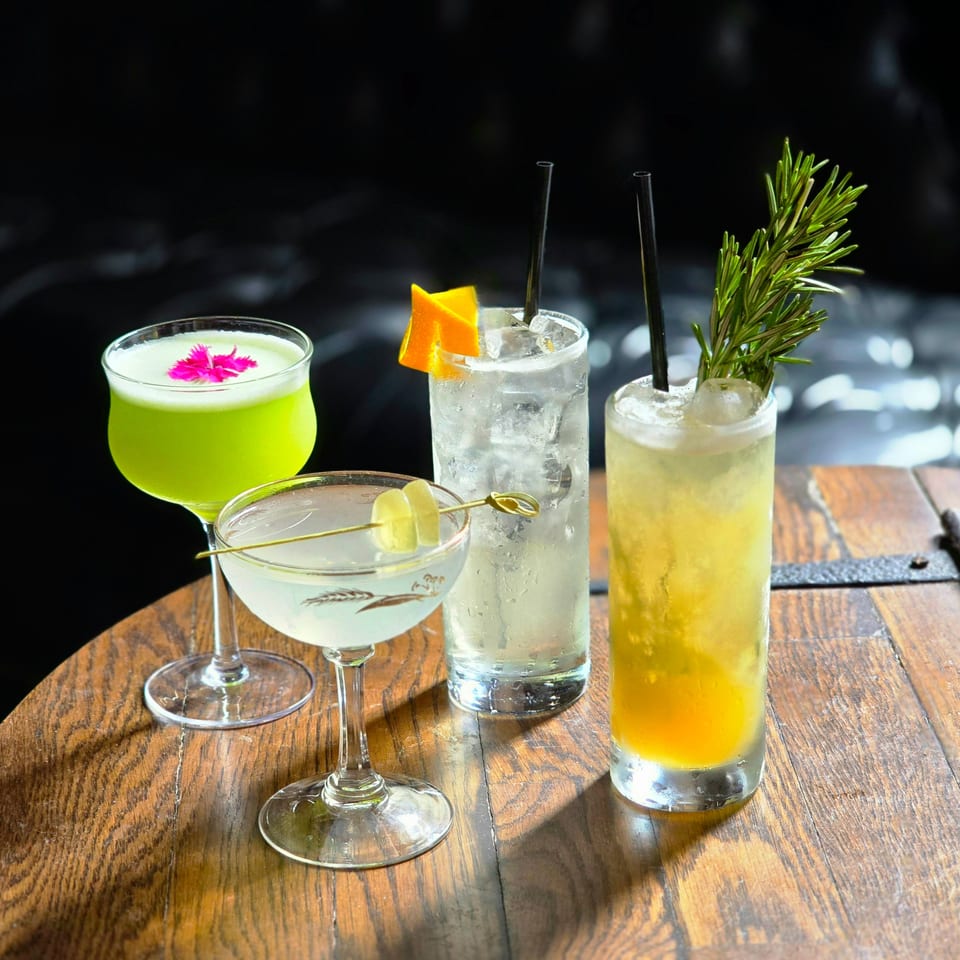 Assorted colorful cocktails with ice, citrus garnishes, edible flower, and rosemary sprigs arranged on a wooden restaurant table.