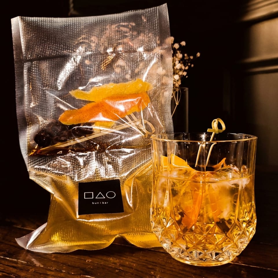 Amber cocktail with citrus garnish beside a branded snack bag on a dimly lit wooden bar.