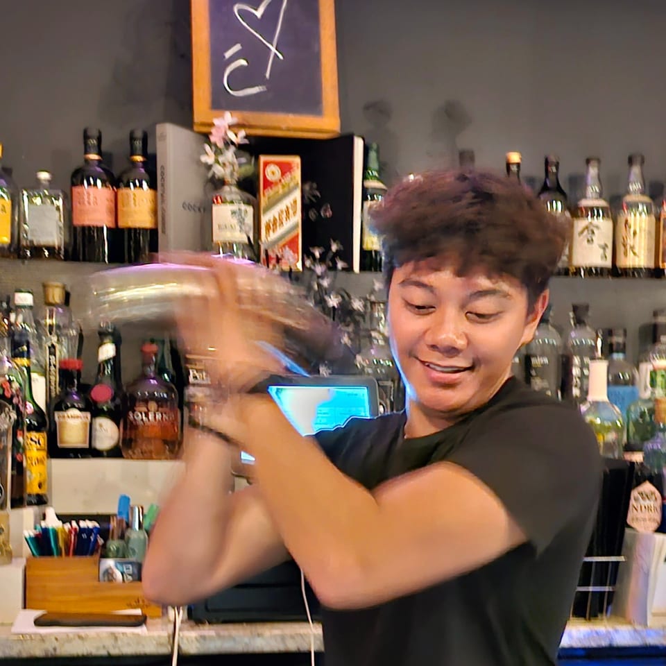 Bartender smiles while mixing a drink behind a bar stocked with liquor bottles and illuminated by a purple neon sign.