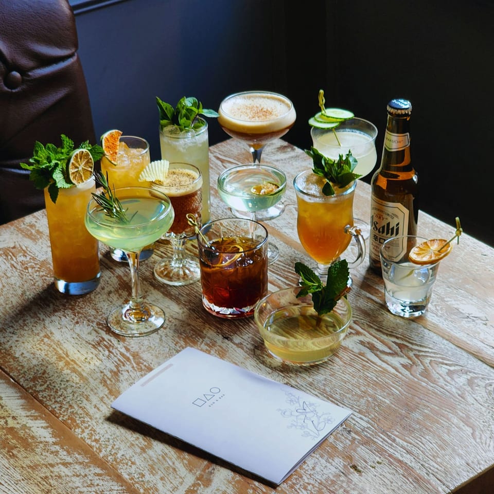 Assorted colorful cocktails garnished with citrus, herbs, cucumber, and flowers surround a Salhi beer bottle on a rustic restaurant table.