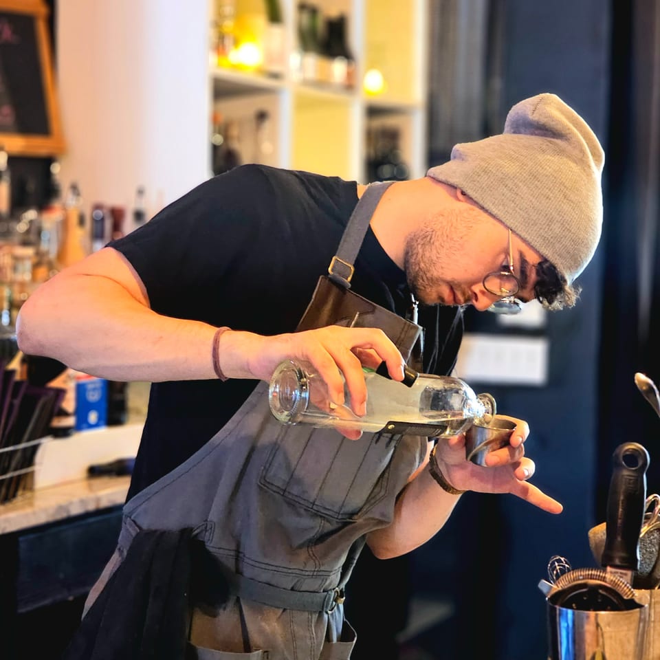 Bartender in a beanie and apron pours liquor from a bottle into a jigger at a warmly lit bar.