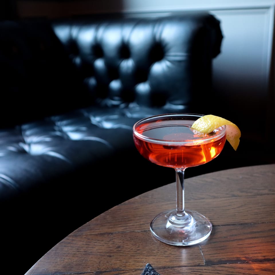 Red cocktail in a coupe glass garnished with a lemon twist, displayed on a wooden table in a dimly lit restaurant.