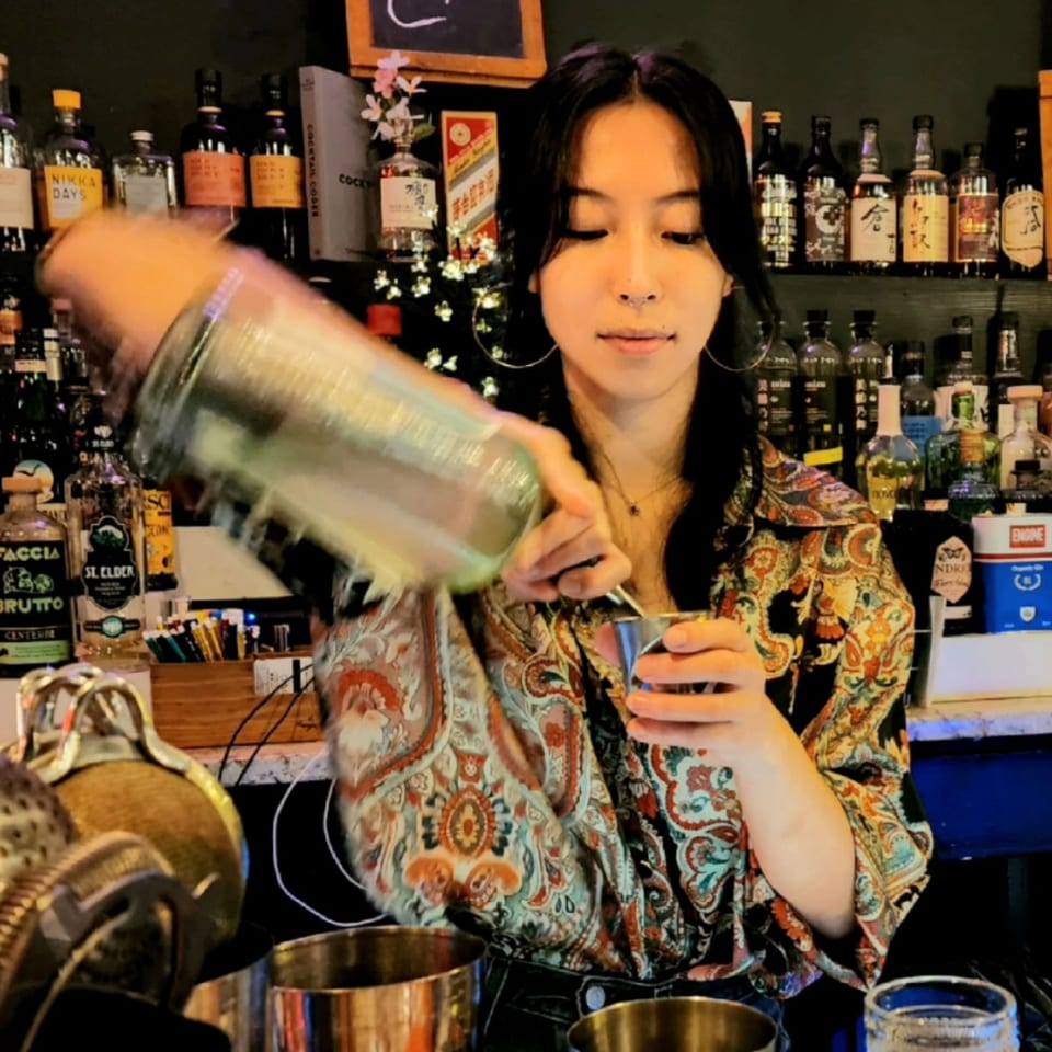 Bartender shakes a cocktail in a metal shaker behind a bar lined with liquor bottles.