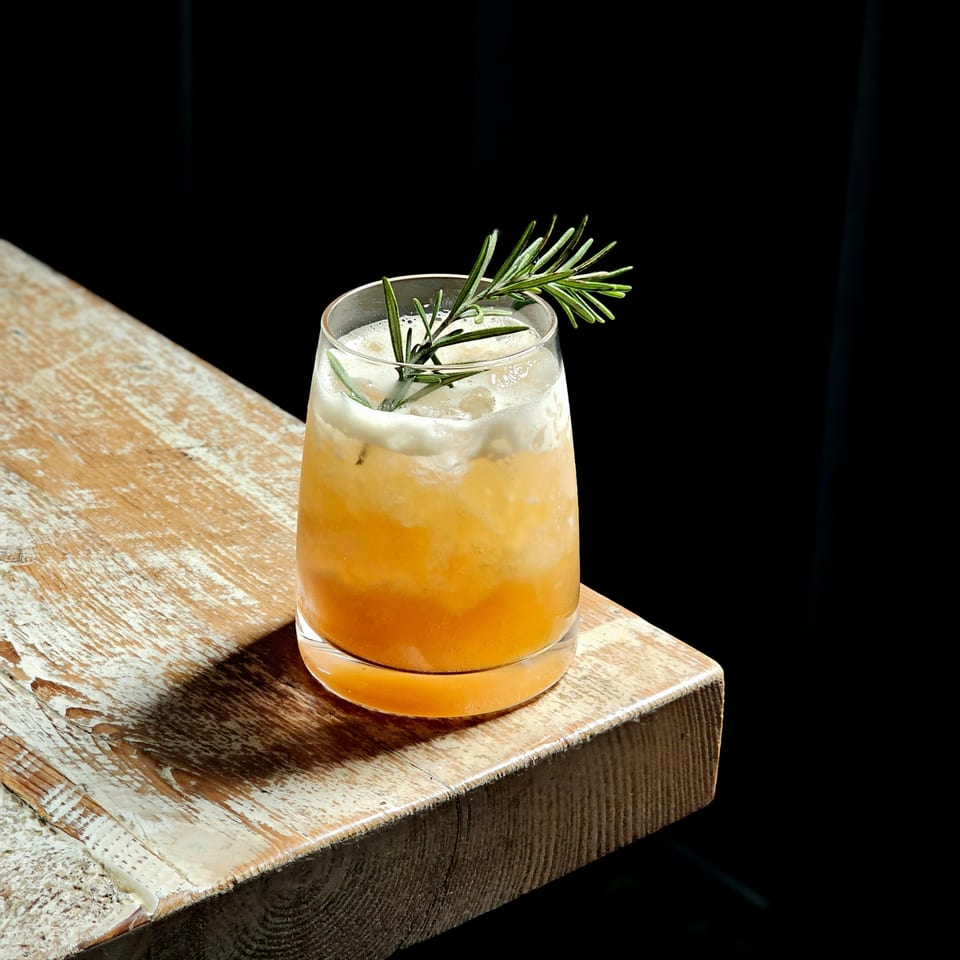 Amber iced cocktail topped with foam and a fresh rosemary sprig, resting on a rustic wooden table.