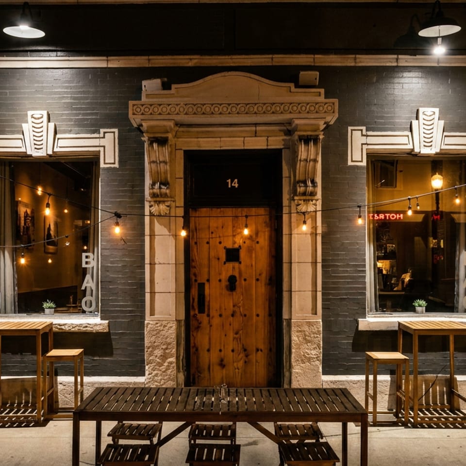Nighttime view of a warmly lit restaurant facade with ornate stonework, wooden door, outdoor tables, and string lights.