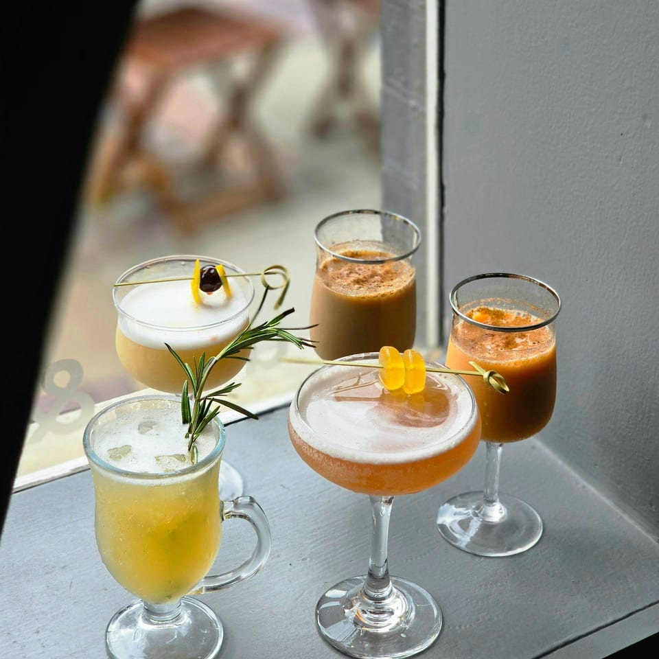 Assorted colorful cocktails in elegant glasses, garnished with citrus, cherries, and rosemary on a sunlit restaurant table.